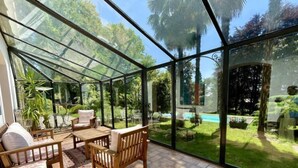 Ein Wintergarten mit Glasdach, Holzmöbeln und Blick auf Pool und Garten.
