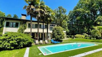 Une maison à deux étages avec une piscine, entourée d’une végétation luxuriante et de palmiers.