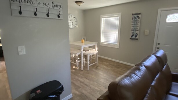 A living room with a brown leather couch, a dining area with a white table and chairs, a wall-mounted clock, and a 'welcome to our home' sign. - 3.2 miles to the beaches and Bay, Cute Florida Cottage (Fort Walton Beach)