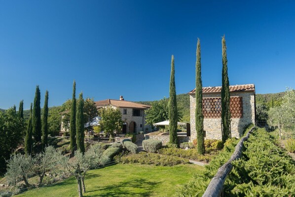 A stone building with a tiled roof, surrounded by a well-maintained garden with tall cypress trees and olive trees.