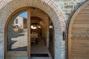 Una entrada de piedra en arco que conduce a un comedor con una mesa lista para dos, una puerta de madera a la derecha y un edificio de piedra al fondo.