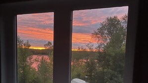 Finestra amb vista a un capvespre sobre un llac, envoltat d'arbres i un tonell de fusta.