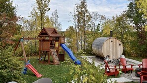 Una caseta de joc de fusta amb tobogan, sauna i cadires vermelles en un jardí.