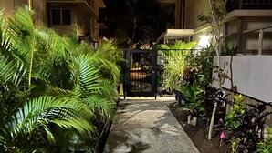Vista nocturna de un edificio residencial con entrada protegida por un portón, rodeado de abundante vegetación y un jardín bien cuidado.