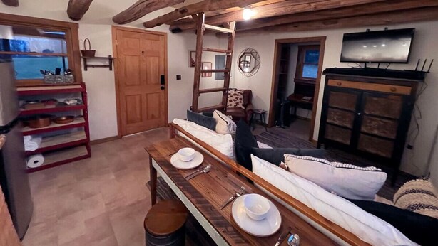 A rustic interior with exposed wooden beams, a dining table set with plates and cutlery, a sofa, and a television mounted on the wall. - Small cabin on private 400 acre mountaintop farm! (Hinton)