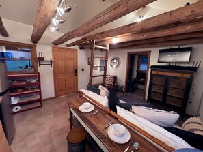 A rustic interior with exposed wooden beams, a dining table set with plates and cutlery, a sofa, and a television mounted on the wall. - Small cabin on private 400 acre mountaintop farm! (Hinton)