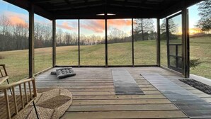 A covered outdoor area with a wooden deck, a cushioned seating area, and a view of a grassy field. - Small cabin on private 400 acre mountaintop farm! (Hinton)