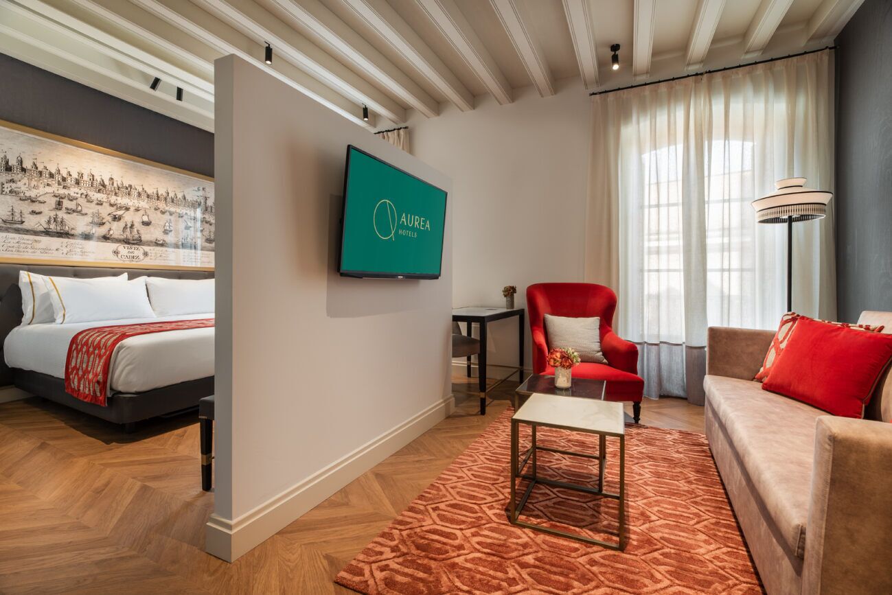 Una habitación de hotel con una cama, un sillón rojo, un sofá, una mesa de centro y un televisor montado en la pared.