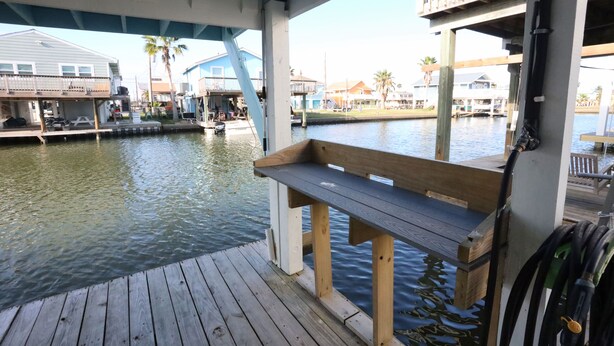 A wooden dock with a bench, a boat, and a waterway with houses in the background.