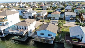 A residential area with houses connected by walkways over water, featuring a mix of single and multi-story buildings with balconies and outdoor seating areas.