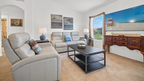 Una sala de estar con un sofá beige, una mesa de centro negra y vista a la playa a través de un ventanal amplio.