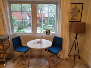 Enjoy a view of the outdoors from the large window, featuring a red building and lush greenery. The room includes a small table, two chairs, and a floor lamp.