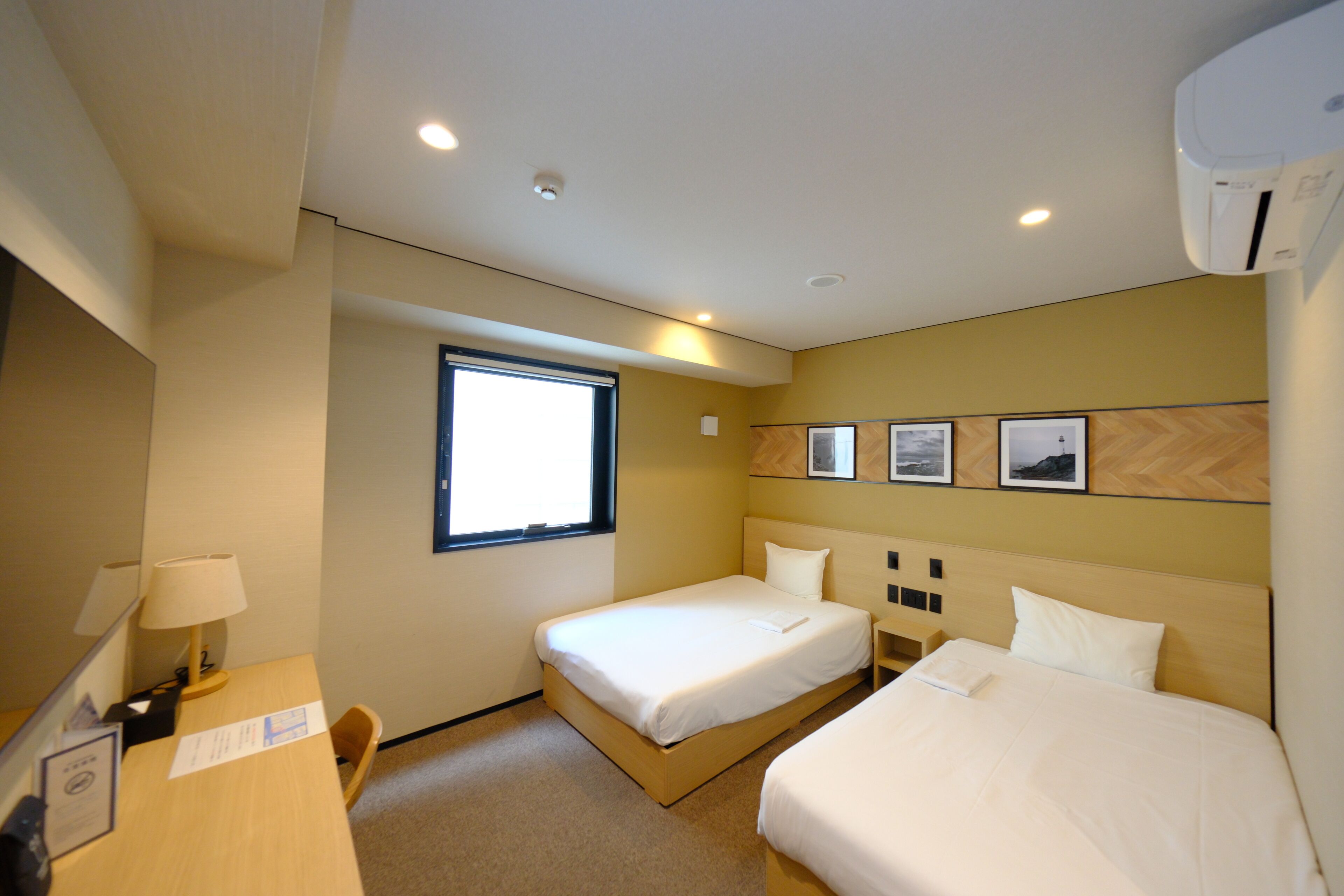 Une chambre d’hôtel avec deux lits, un bureau, une chaise, une fenêtre et un climatiseur fixé au mur.