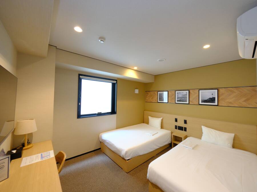 Une chambre d’hôtel avec deux lits, un bureau, une chaise, une fenêtre et un climatiseur fixé au mur.