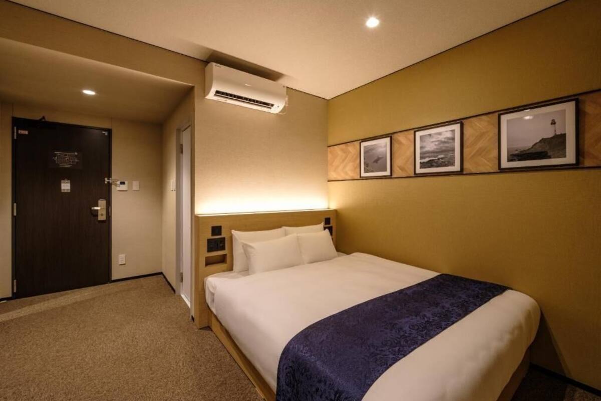 A hotel room with a bed, a wall-mounted air conditioner, and framed pictures on the wall.