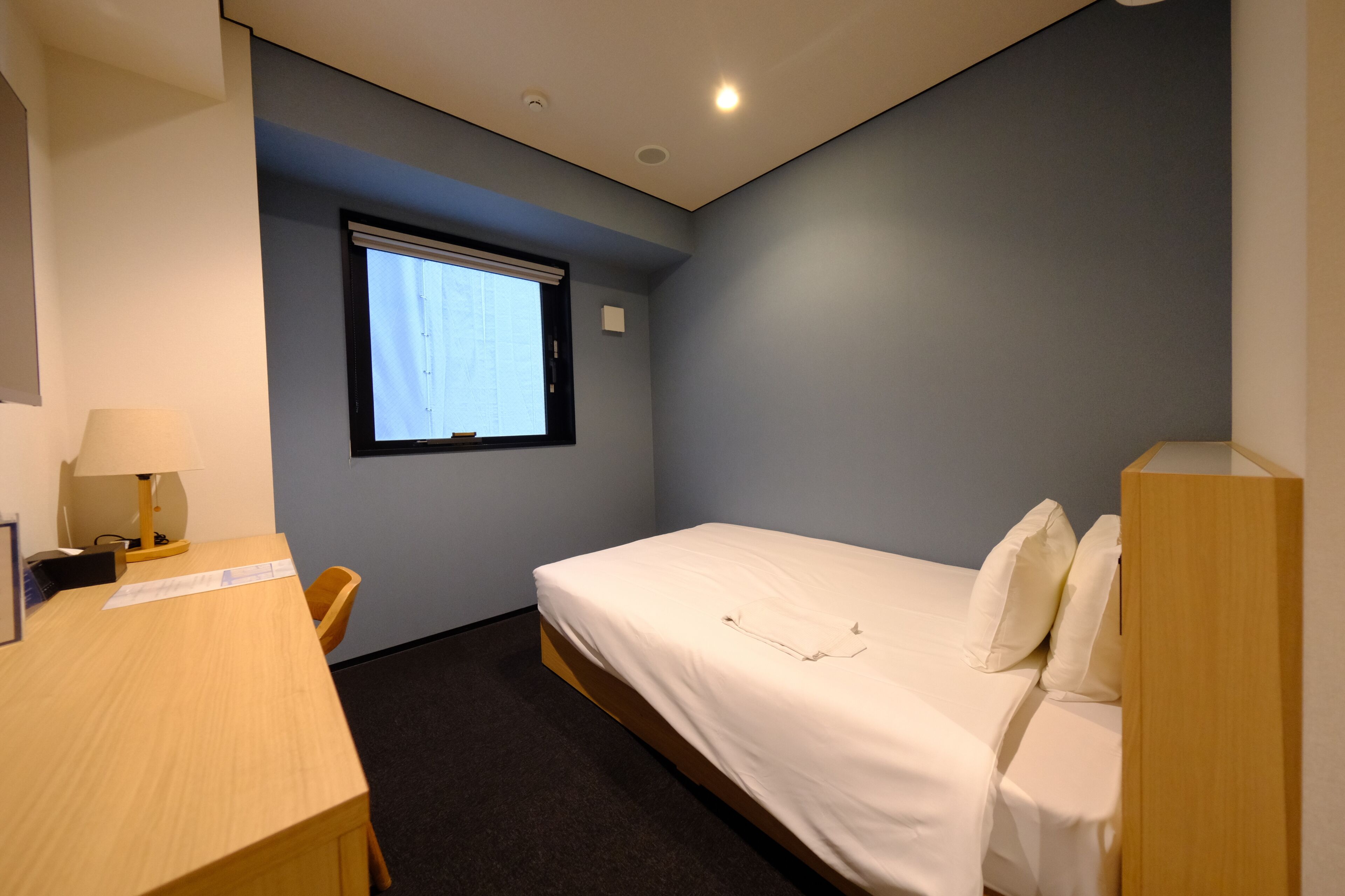 A small, well-lit room with a single bed, a desk, and a chair.