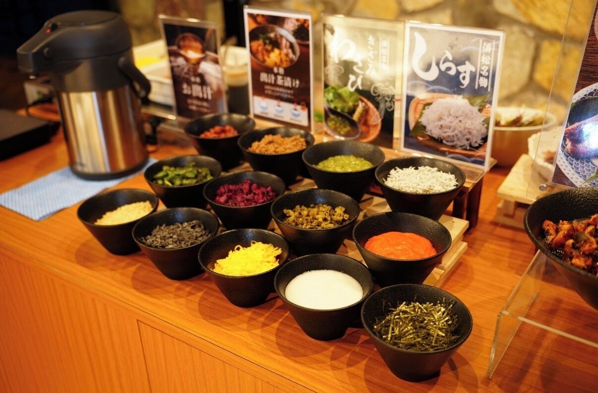 Un bufé con diferentes alimentos en recipientes negros, una cafetera de acero inoxidable y carteles informativos.