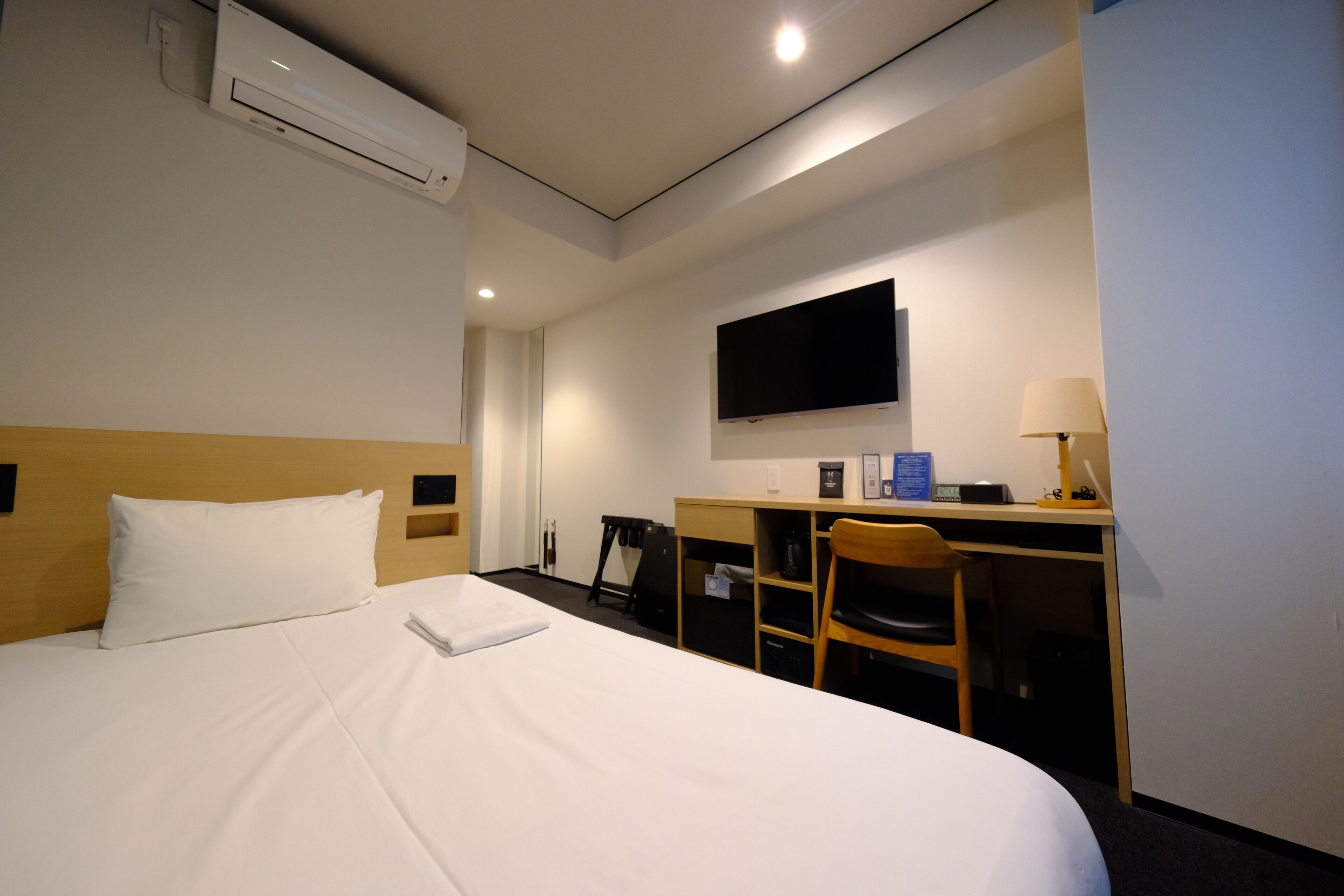 A hotel room with a bed, a desk, a chair, a TV, and an air conditioning unit.