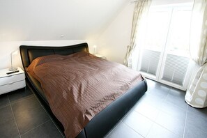 A bedroom with a bed, nightstand, and a window with curtains. - Villa Mare Apartment 4 - Villa Mare (Timmendorfer Strand)