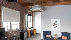 Un espacio de oficina moderno con pared de ladrillo, ventanas amplias, un escritorio, dos sillones, un televisor y un equipo de aire acondicionado.