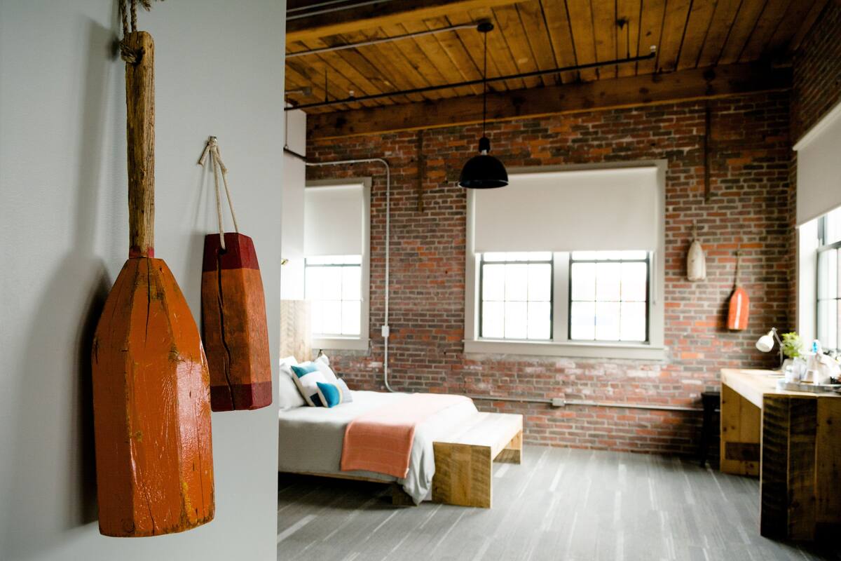 Ein Zimmer mit Backsteinwand, Holzbalken, einem Bett, einem Schreibtisch und einer Pendelleuchte.
