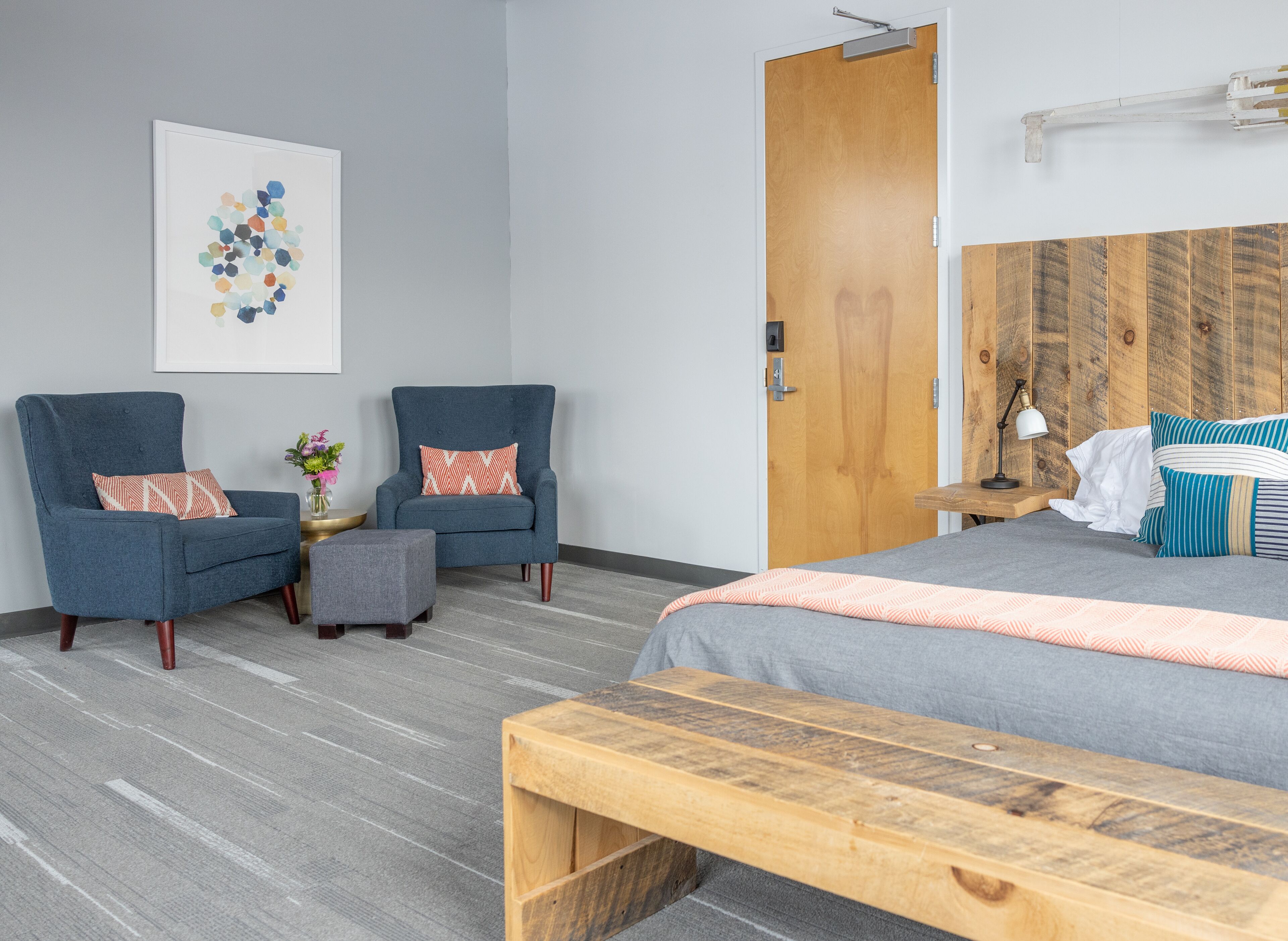 A modern hotel room with a grey sofa, a wooden bench, a bed with striped pillows, and a wooden headboard.