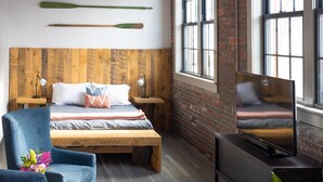 Un dormitorio con cabecera de madera, una cama con almohadas, un sillón azul, un pequeño otoman, un televisor sobre un mueble bajo y una planta en maceta.