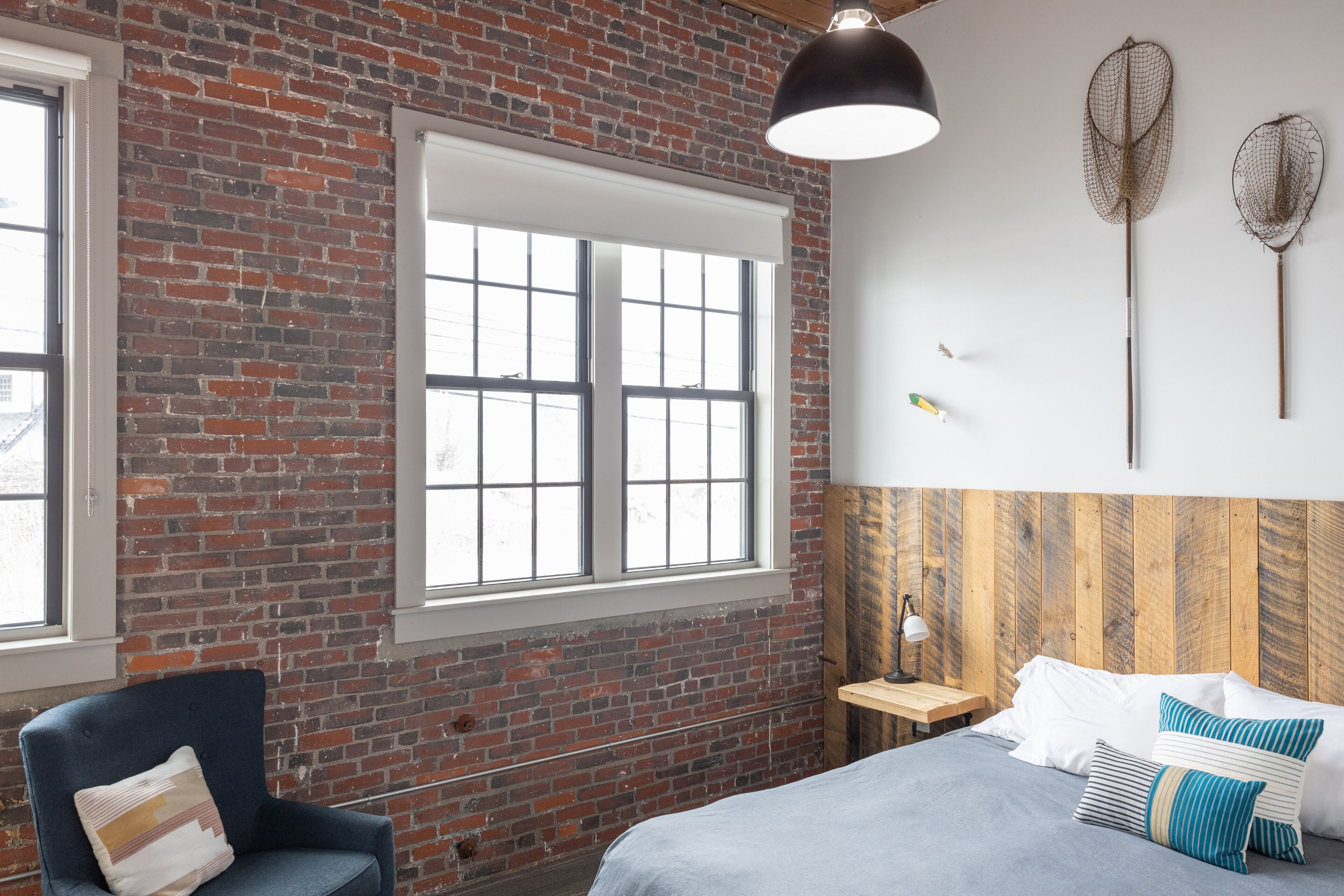 A room with a brick wall, a large window, a bed with pillows, a chair, and two wall-mounted nets.