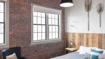 A room with a brick wall, a large window, a bed with pillows, a chair, and two wall-mounted nets.