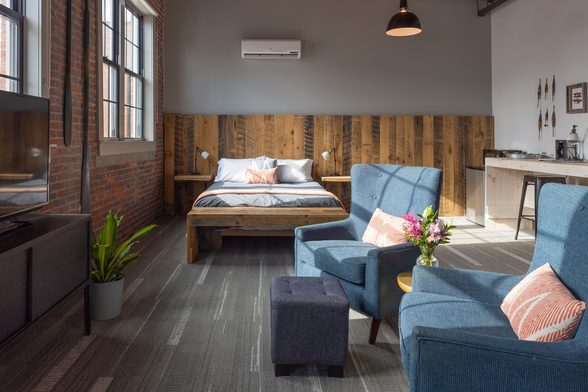 Ein modernes Hotelzimmer mit einem großen Bett, einer Sitzecke mit einer Couch und Sesseln, einem kleinen Tisch sowie einer Küchenzeile.