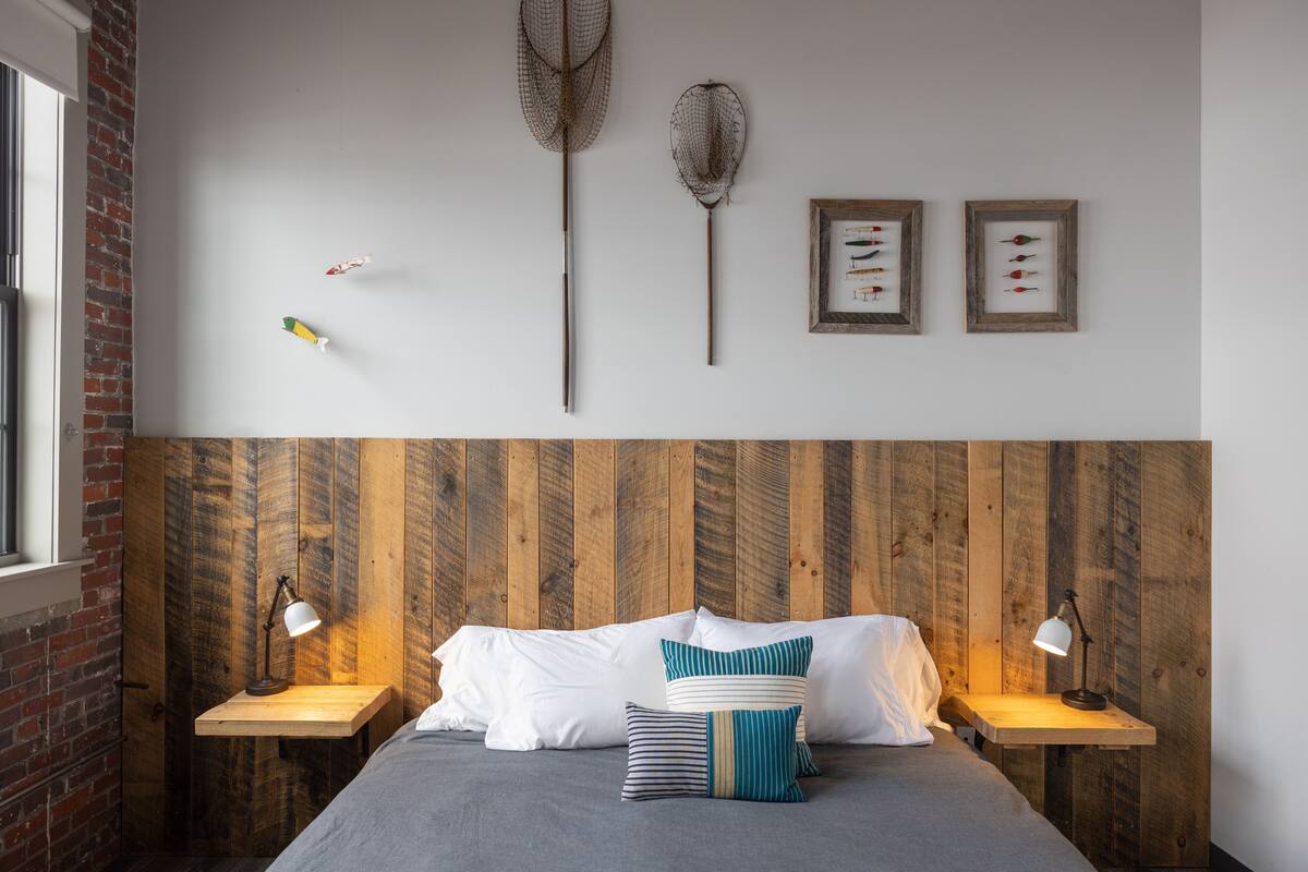 Ein Schlafzimmer mit einem Holzkopfteil, zwei Nachttischen mit Lampen, einem Bett mit weiß gestreiften Kissen sowie an den Wänden befestigten Fischernetzen und gerahmten Fischabbildungen.