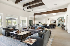 A spacious living area with a large sofa, a coffee table, and a dining table. There is a kitchen in the background with modern appliances.