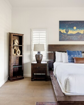 A bedroom with a bed, a nightstand with a lamp, a wooden cabinet with decorative items, and a painting of a desert landscape.