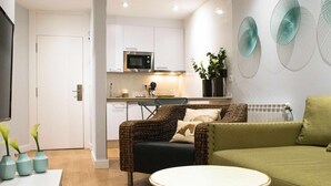 Un soggiorno moderno con un divano verde, un tavolino rotondo e una poltrona in vimini. Si intravede sullo sfondo una cucina con mobili bianchi e un forno a microonde.