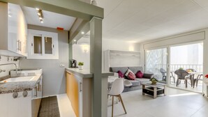 En modern lägenhet med kök, matplats och vardagsrum. Den har ett stort fönster, en soffa och ett soffbord.