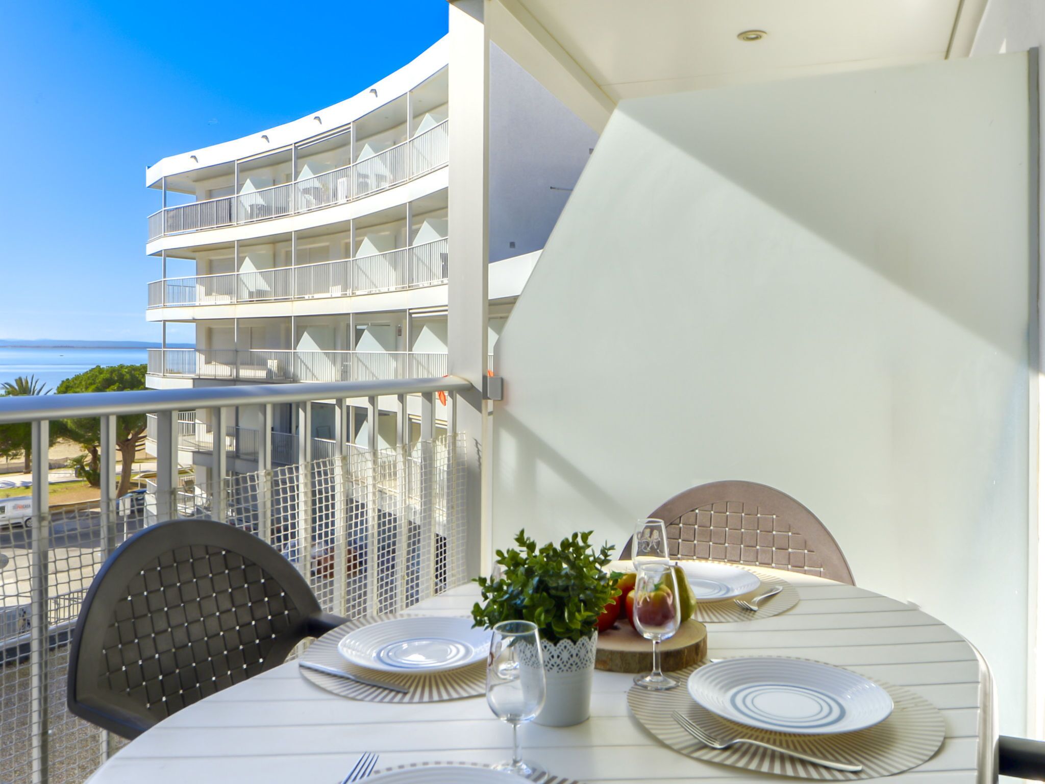 A dining table set with plates, cutlery, and a glass of water on a balcony overlooking a building with multiple balconies and a clear blue sky.