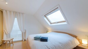 Un dormitorio con tragaluz, cama con ropa blanca, mesita de noche de madera, lámpara de noche, una silla y ventana con cortinas blancas.