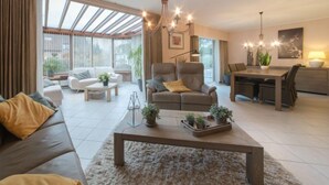 Amplia sala de estar con sofá, sillón y mesa de centro, zona de comedor con mesa y sillas, y ventanas grandes que permiten la entrada de luz natural.