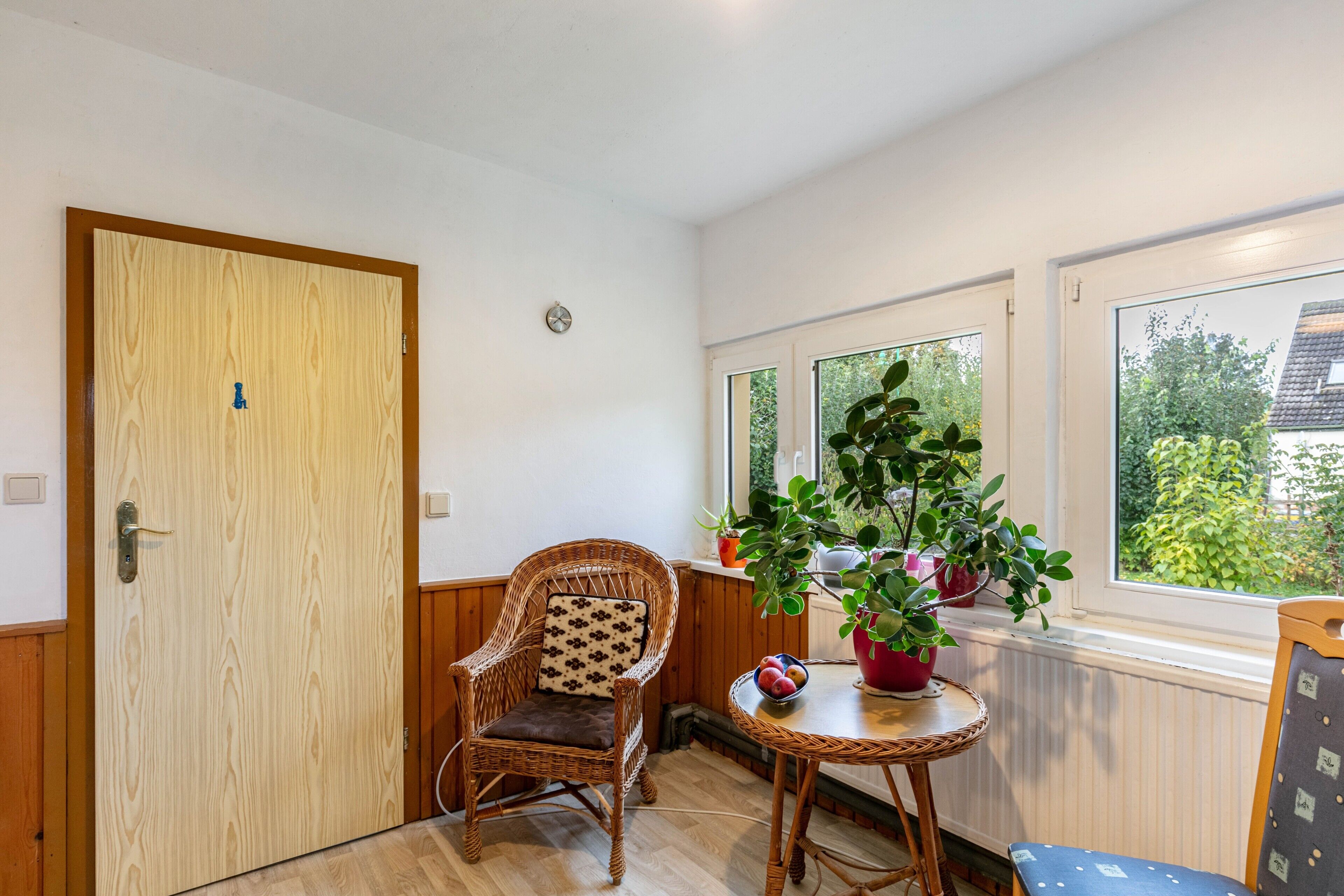 Une pièce avec une porte en bois, une chaise en osier dotée d’un coussin, une petite table ronde sur laquelle est posée une plante en pot, et une fenêtre donnant sur un paysage verdoyant.
