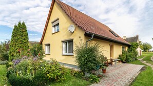 Une maison jaune avec un toit rouge, portant les numéros 16 et 18, une antenne parabolique et un jardin où poussent diverses plantes et fleurs.