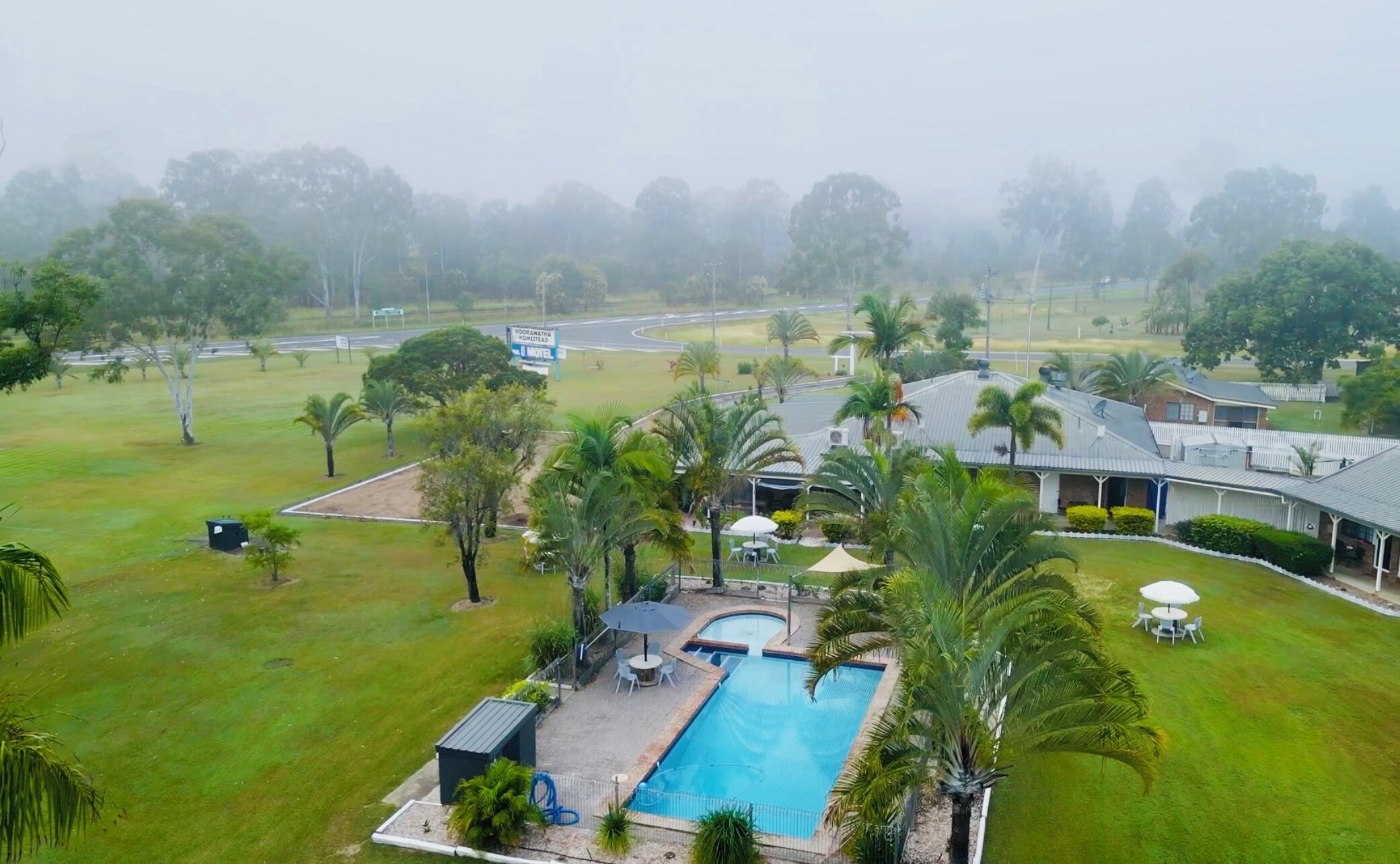 A hotel with a swimming pool, outdoor seating, and a view of a road and trees.