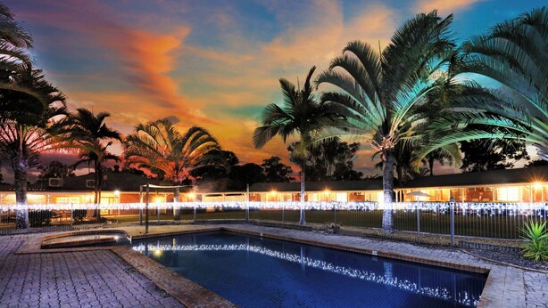 Zona de piscina con palmeras, un edificio iluminado y un cielo al atardecer.