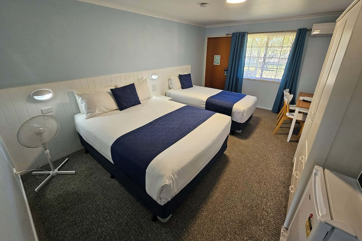 Een hotelkamer met twee bedden, een bureau, een stoel, een ventilator en een raam met gordijnen.