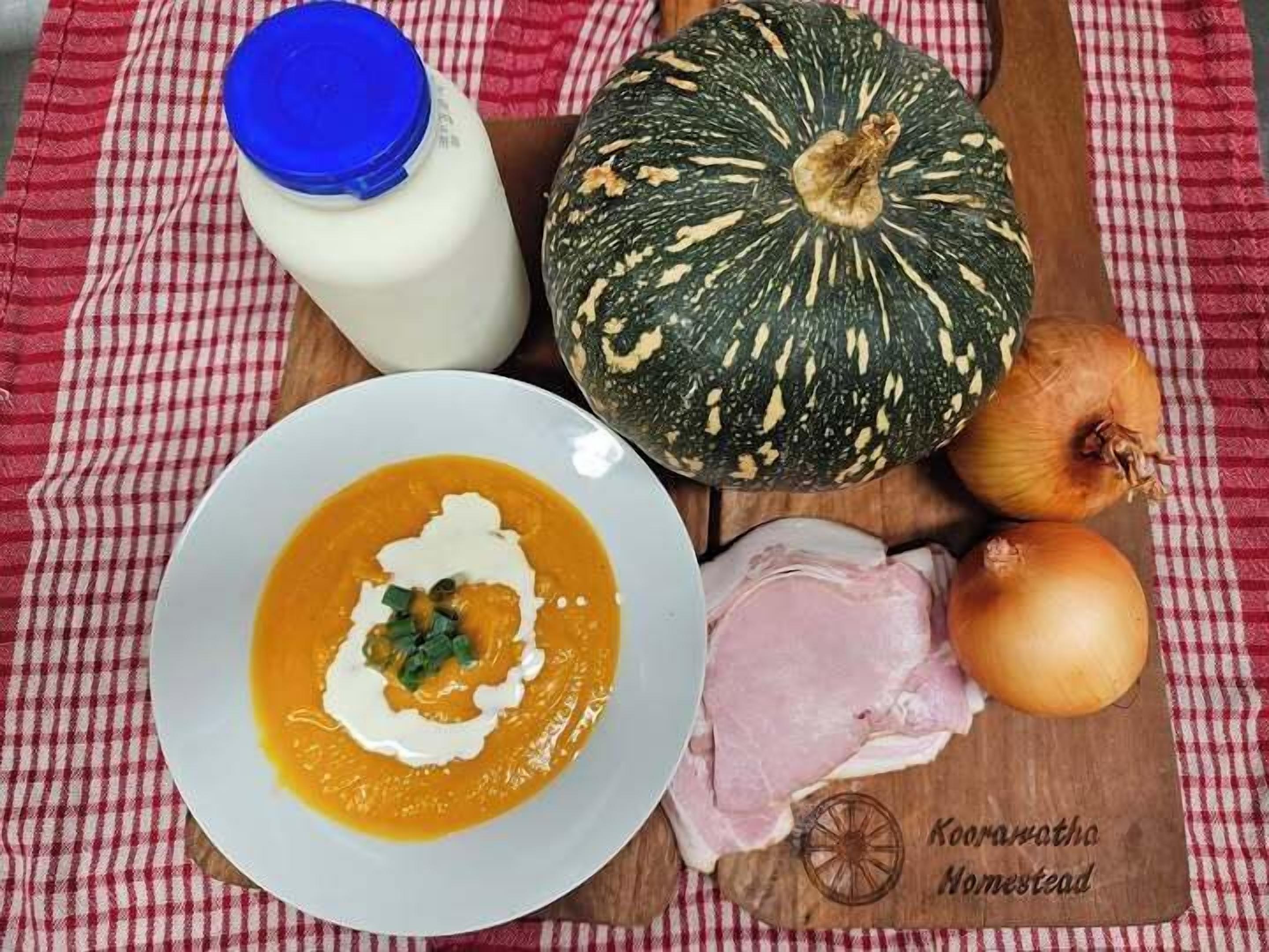 A bowl of soup with a dollop of cream and green garnish, a pumpkin, a bottle of milk, raw meat, and onions on a wooden board with a red and white checkered cloth underneath.