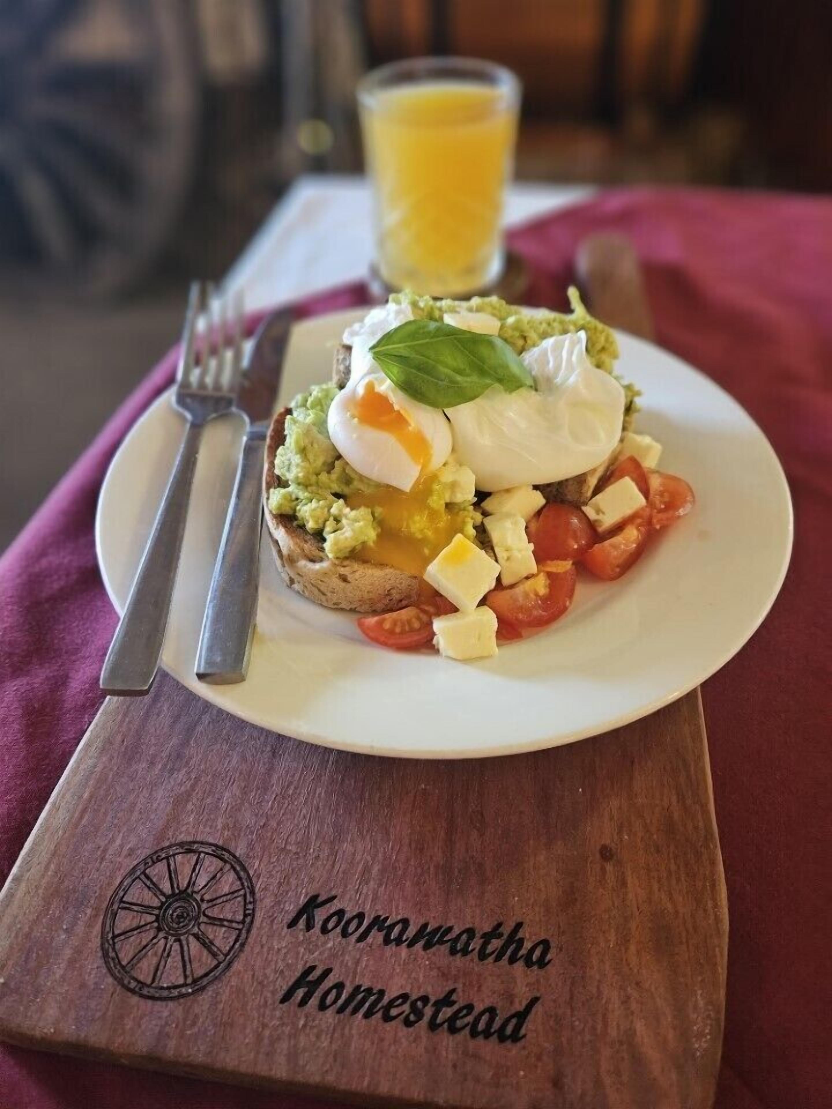 A breakfast plate with avocado toast, a poached egg, and tomatoes on a wooden board labeled "Kooranatha Homestead".