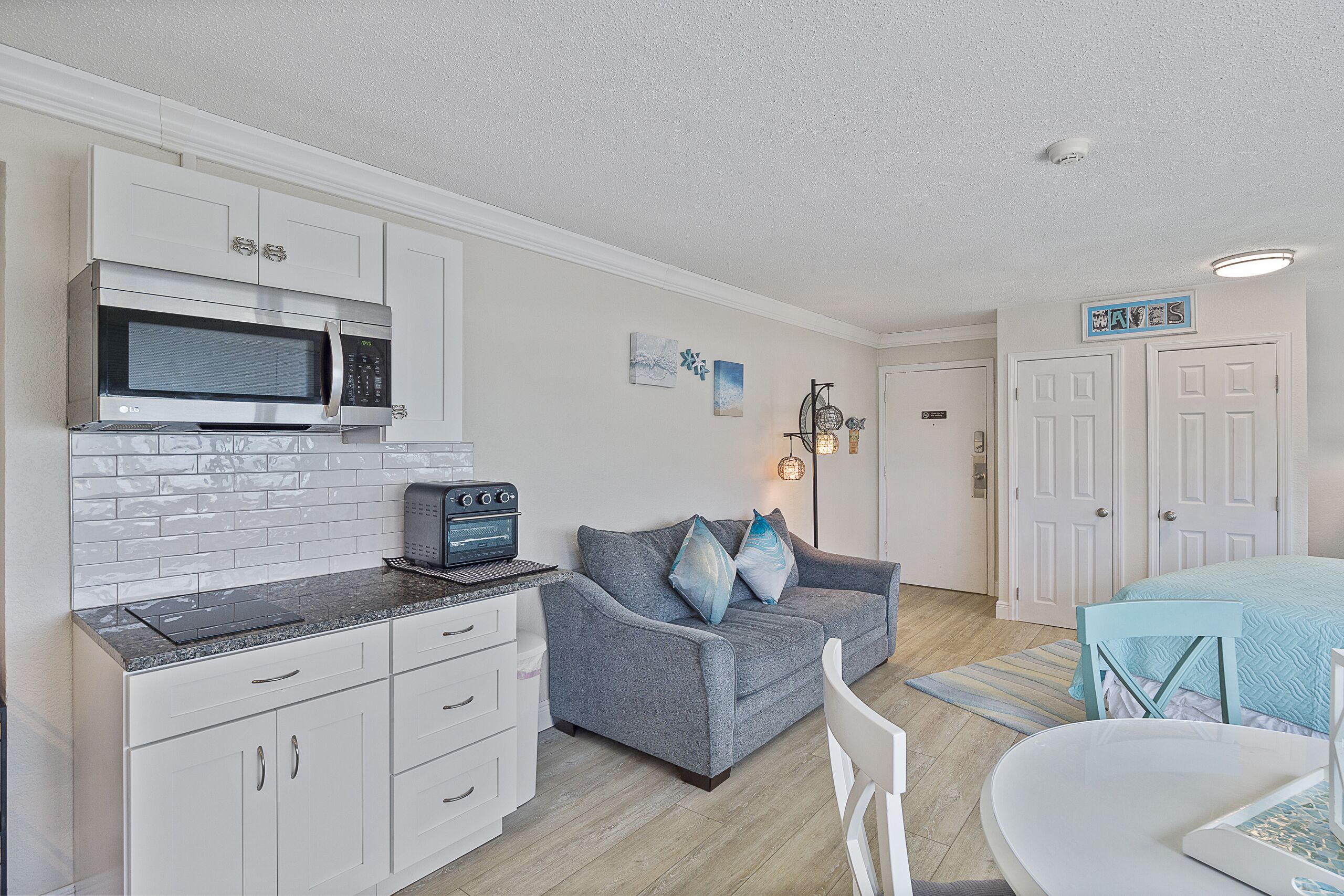 A compact kitchen with a microwave, toaster, and white cabinets. There is a gray sofa with blue pillows and a small dining table with a chair.