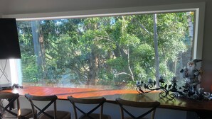 Uma sala de jantar com uma janela ampla que oferece vista para uma floresta densa. Há quatro cadeiras de madeira e uma mesa com um candelabro decorativo.