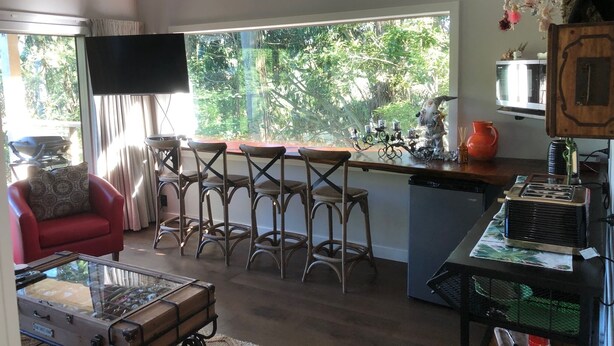 Ampla sala de estar com uma grande janela, um sofá vermelho, uma mesa com tampo de vidro e uma área de cozinha com banquetas.