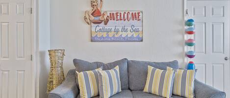 Un sofá gris con almohadones a rayas, un cartel de bienvenida y una guirnalda decorativa de bolas.