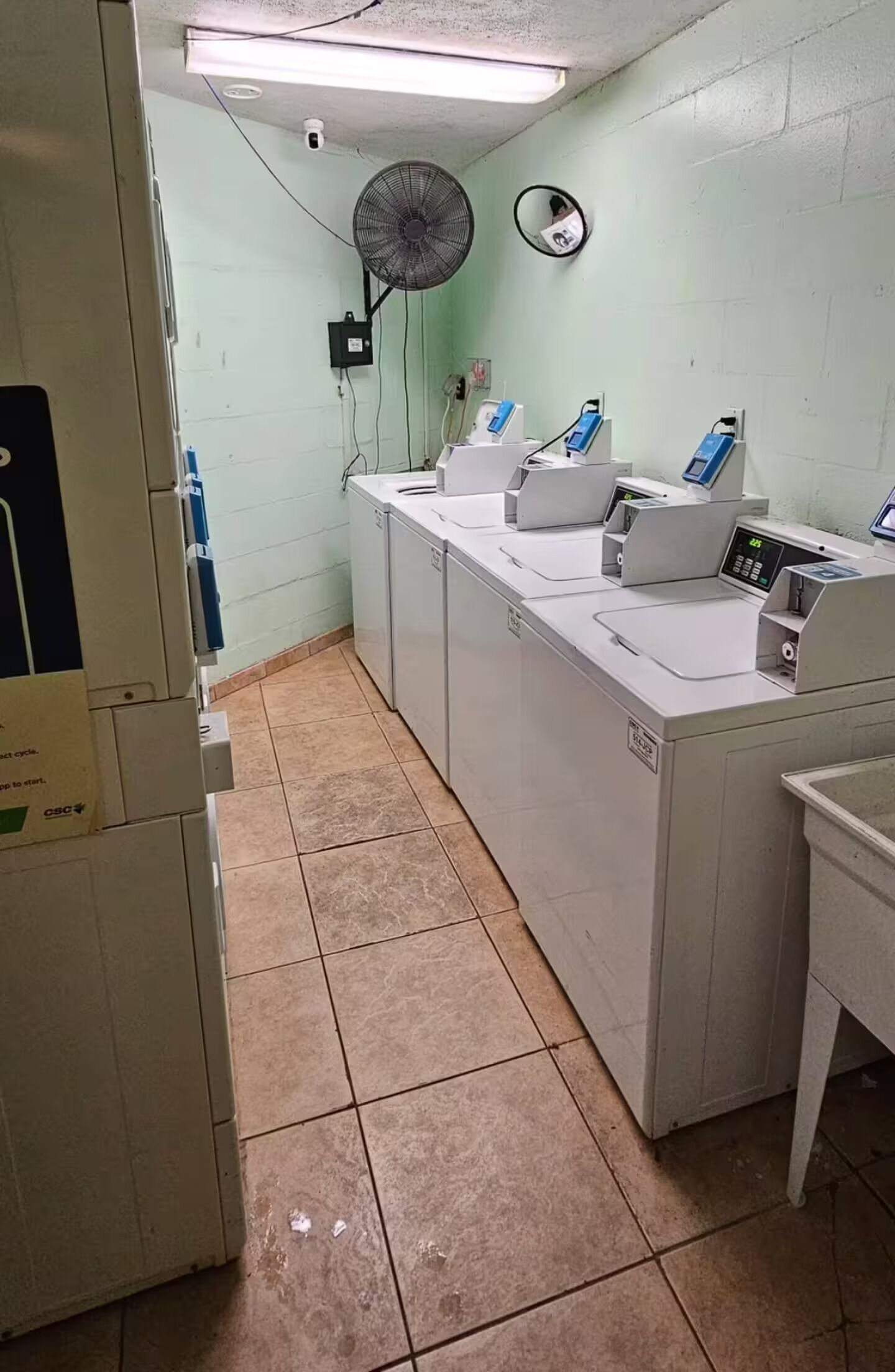 A laundry room with industrial-style washing machines and dryers, a ceiling fan, and a security camera.